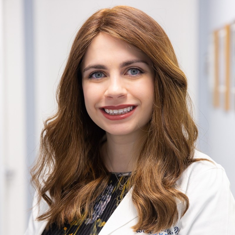 Smiling woman with long auburn wavy hair wearing a black floral blouse and a white lab coat.