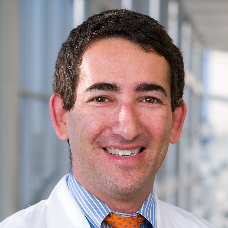 Smiling man with brown hair wearing a light blue dress shirt, yellow tie, and white lab coat