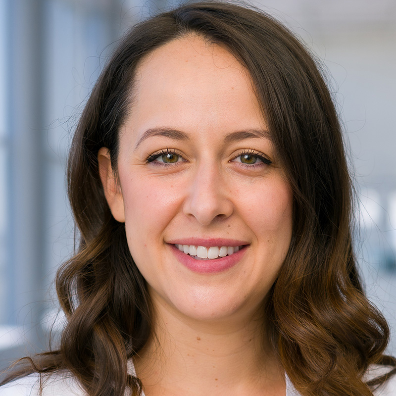 Smiling woman with brown wavy shoulder-length hair wearing a white blouse and white lab coat.