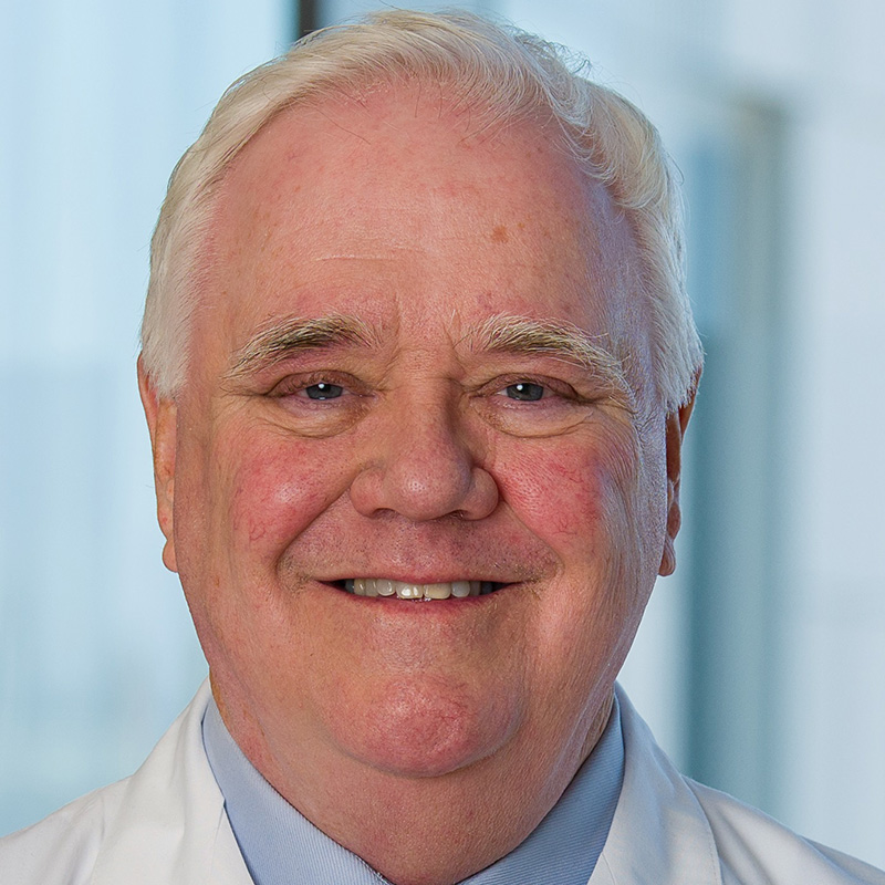 Smiling man with white hair wearing a light blue shirt, blue tie, and white lab coat.