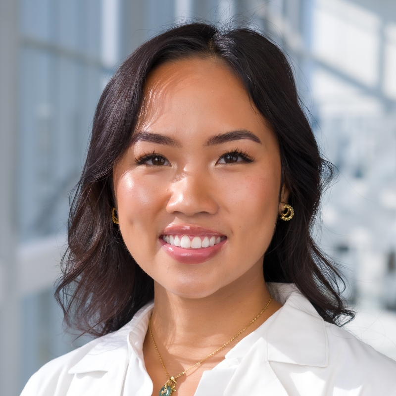 Smiling young woman with wavy, shoulder-length black hair wearing a white blouse, gold jade necklace, gold hoop earrings, and a white lab coat.