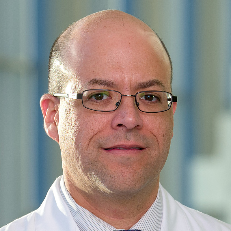 Slightly smiling man with bald head wearing black-framed rectangular glasses, a white and blue striped dress shirt, blue tie, and white lab coat.