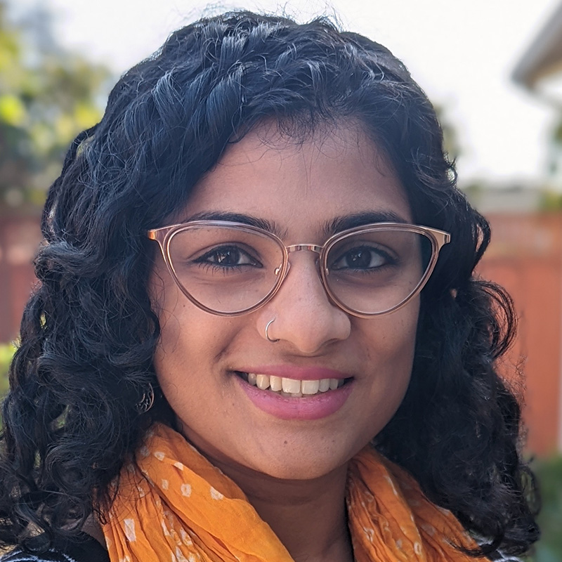 Smiling woman with a nose ring and black curly shoulder-length hair wearing a dark blue and white patterned blouse and a yellow scarf.