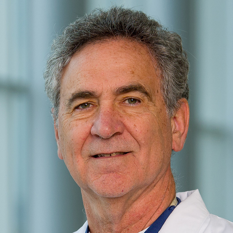 Smiling man with gray hair wearing a white t-shirt, blue scrubs, and white lab coat.