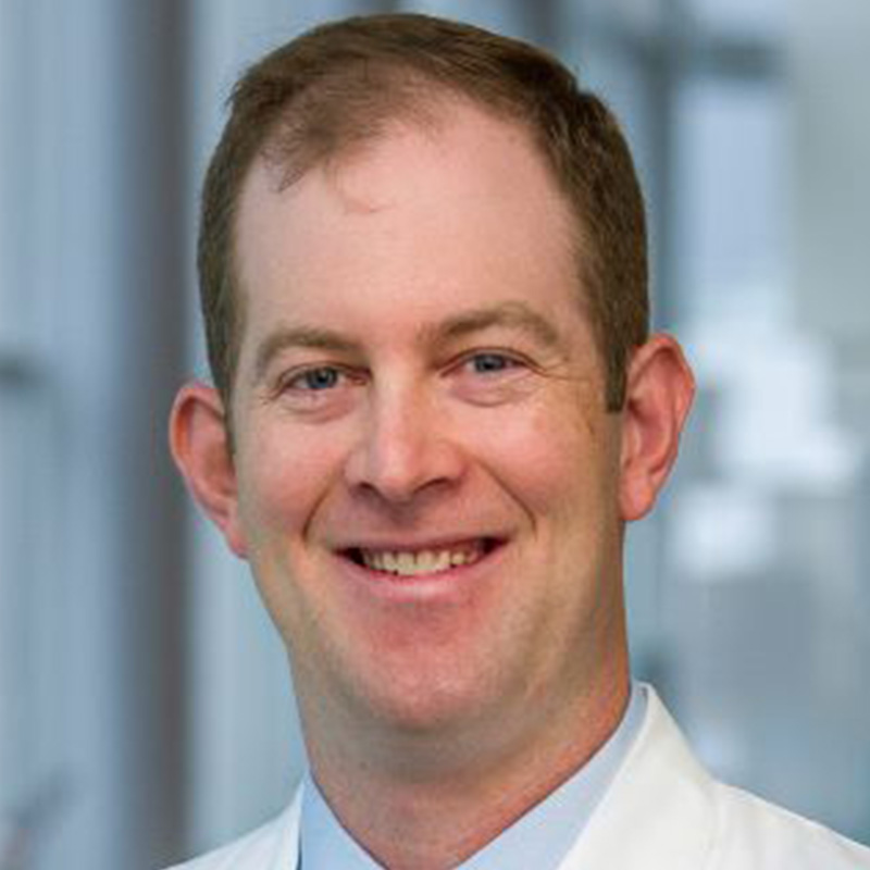 Smiling man with brown hair wearing a light blue dress shirt, dark blue tie and white lab coat.