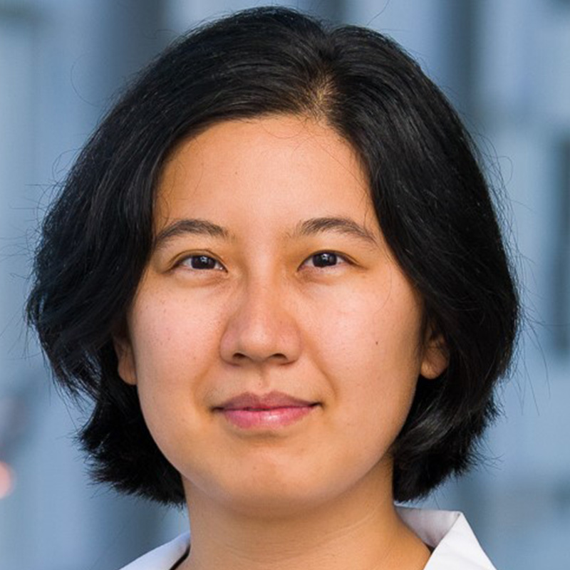 Slighly smiling woman with short black hair wearing a red and black plaid blouse and white lab coat.
