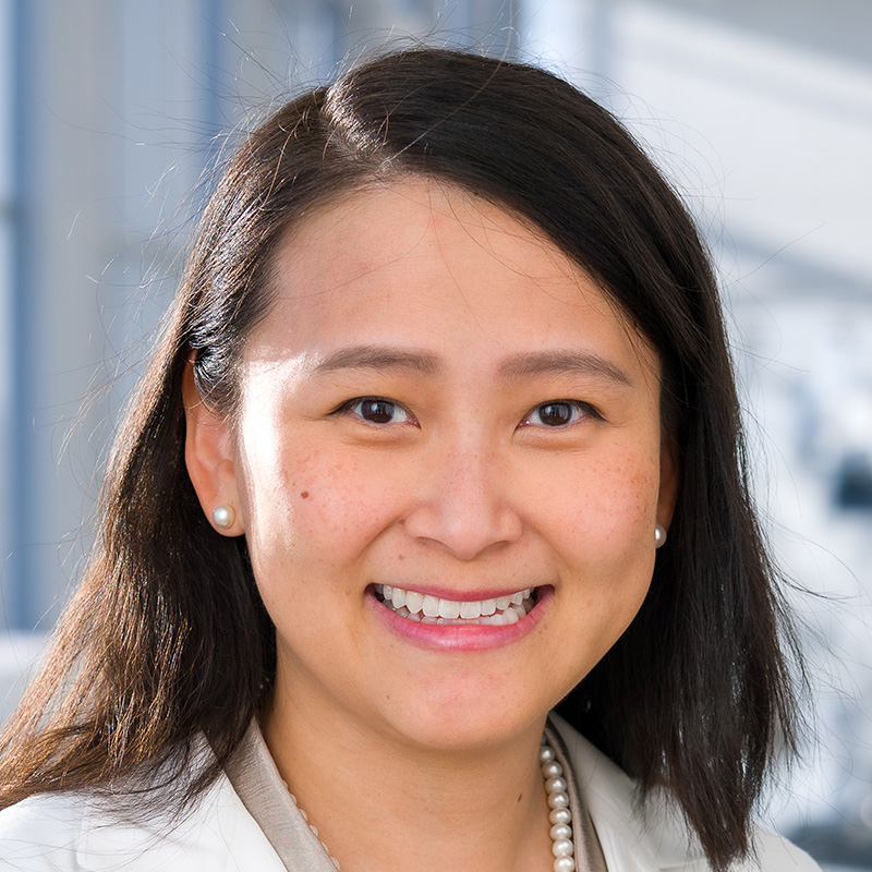 Smiling woman with black shoulder length hair wearing a pearl necklace, champagne-colored blouse, and white lab coat.