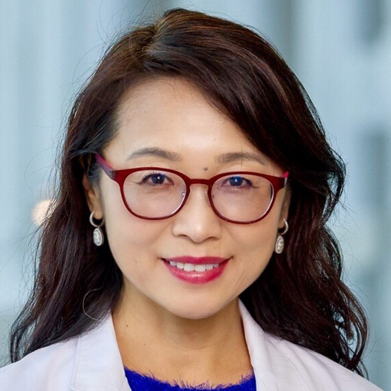 Smiling woman with black wavy hair wearing round red glasses, a fuzzy purple blouse, and white lab coat.