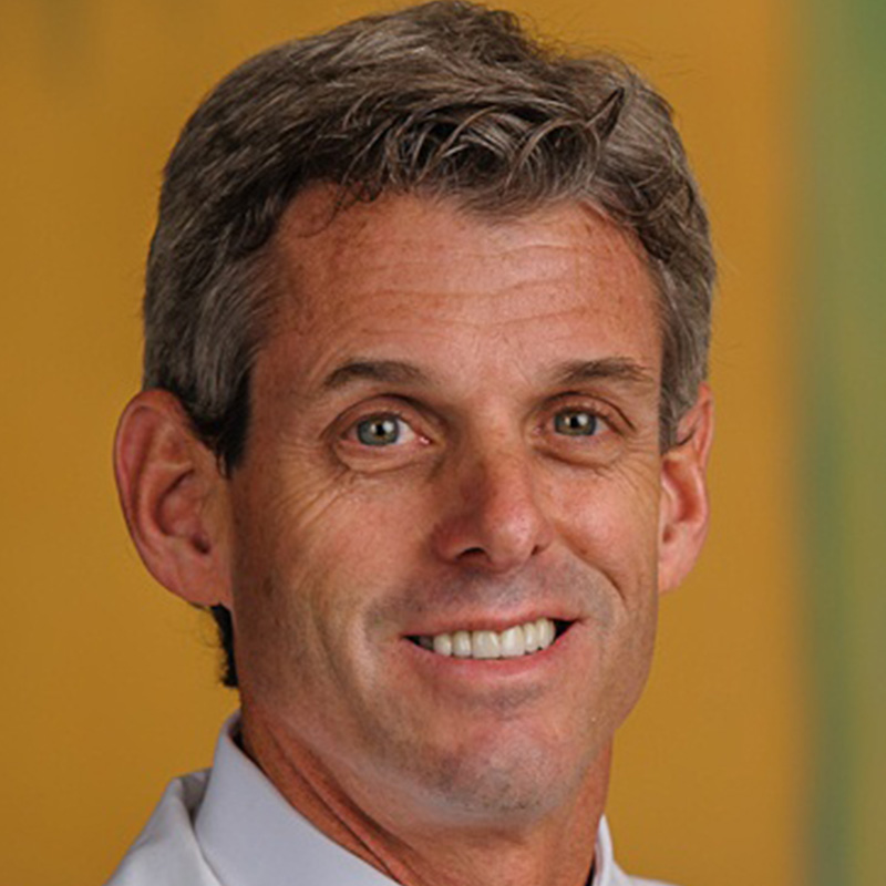 Smiling man with brown-gray hair wearing a white dress shirt, yellow and blue striped tie, and white lab coat.