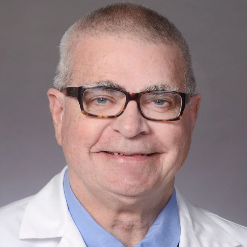 Smiling man with short gray hair wearing thick black-rimmed glasses, a light blue dress shirt, blue tie and white lab coat.