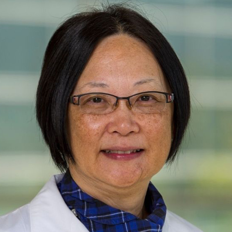 Smiling woman with a black bob wearing black-rimmed rectangular glasses, a checked blue and black blouse, and white lab coat.