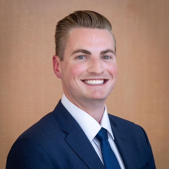 Smiling man with light brown hair wearing a blue suit and tie, with a white dress shirt.