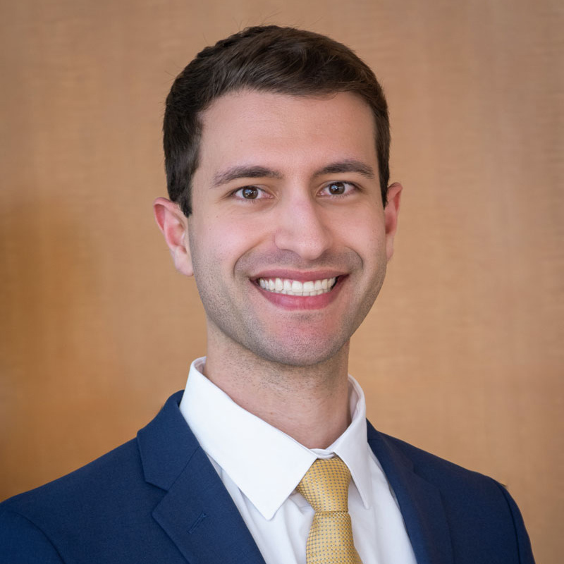 Smiling man with short dark hair, wearing a blue suit, white dress shirt, and gold tie.