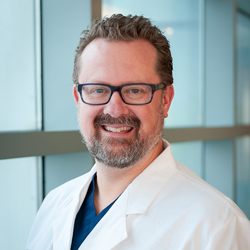 Dr. Sammer, a smiling man with brown hair, mustache and beard, wearing a lab coat and glasses.