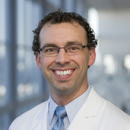 Smiling man with dark curly hair, wearing a lab coat and glasses.
