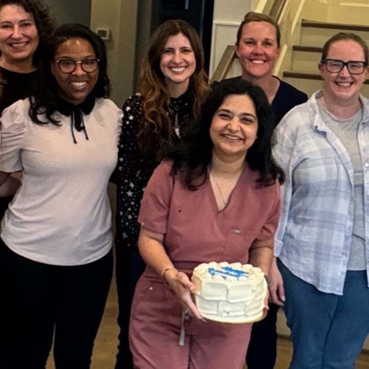 6 people smile as they gather around dark haired woman in the center who is holding a decorated cake