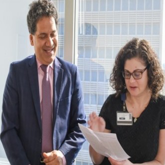 tall white male in blue suit and tie at left smiles at short dark-haired woman gesturing to notes on paper