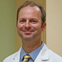 headshot of smiling white man wearing a white lab coat with a blue shirt and a yellow tie