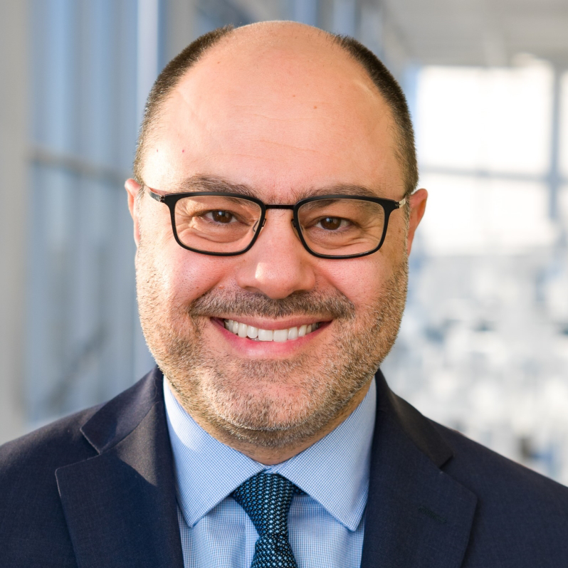 Dr. Tarek Rajji headshot of smiling white male with balding hair, short dark beard wearing glasses and dark jacket with light blue shirt and dark blue tie