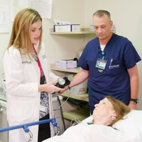 female doctor with long blonde hair in white coat at left, male technician in blue scrubs stand over female patient lying on hospital bed