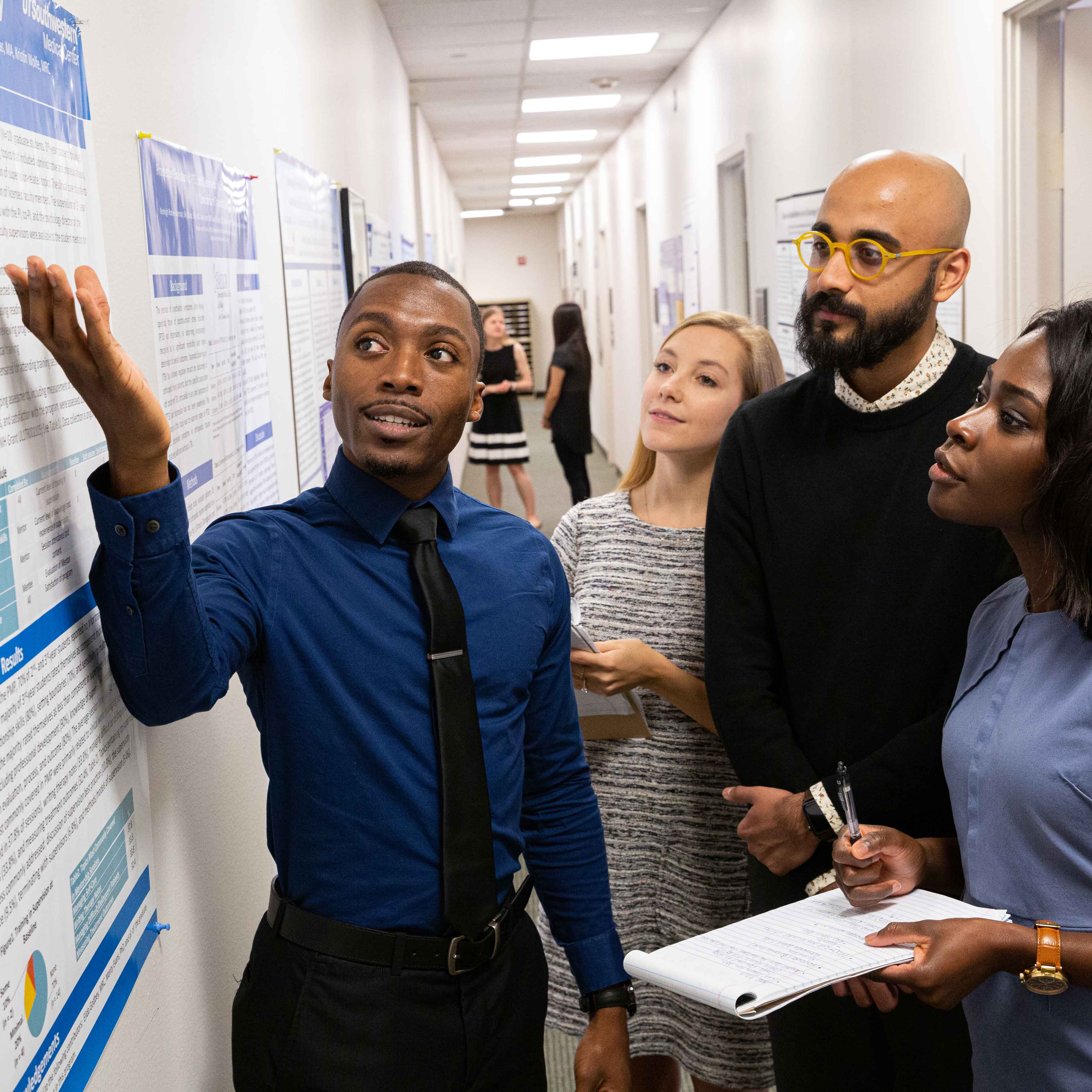 young black man in blue shirt and tie gestures to posters on wall as 2 women and 1 man look on and take notes