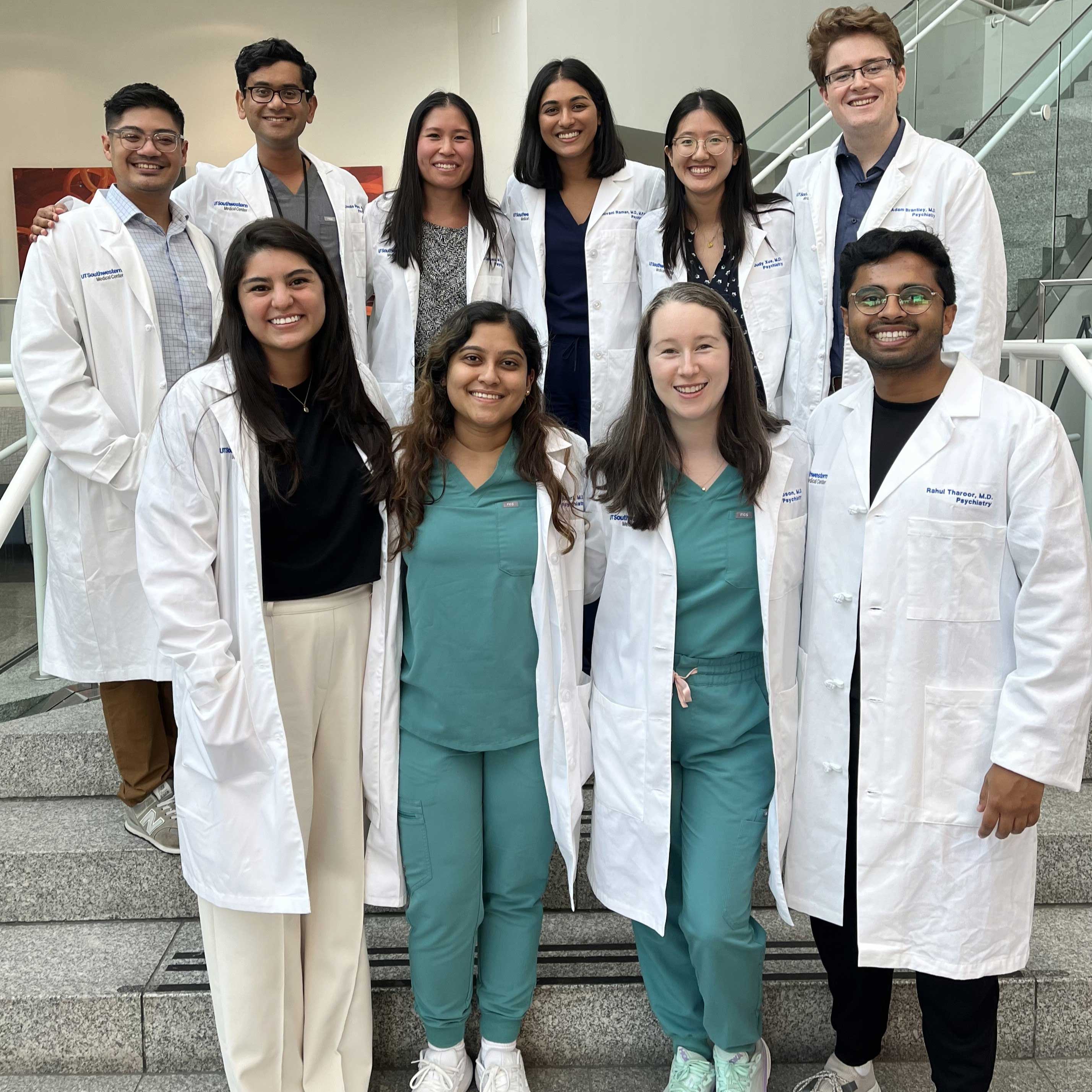 group of 10 young medical students stand on steps wearing green scrubs and white medical coats