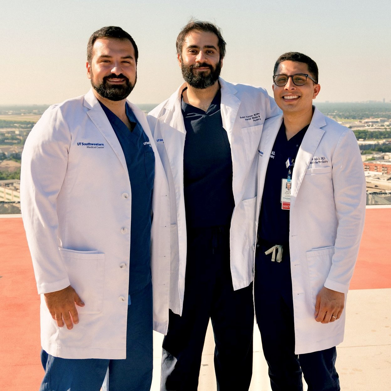 three men stand together wearing blue scrubs and white medical coats