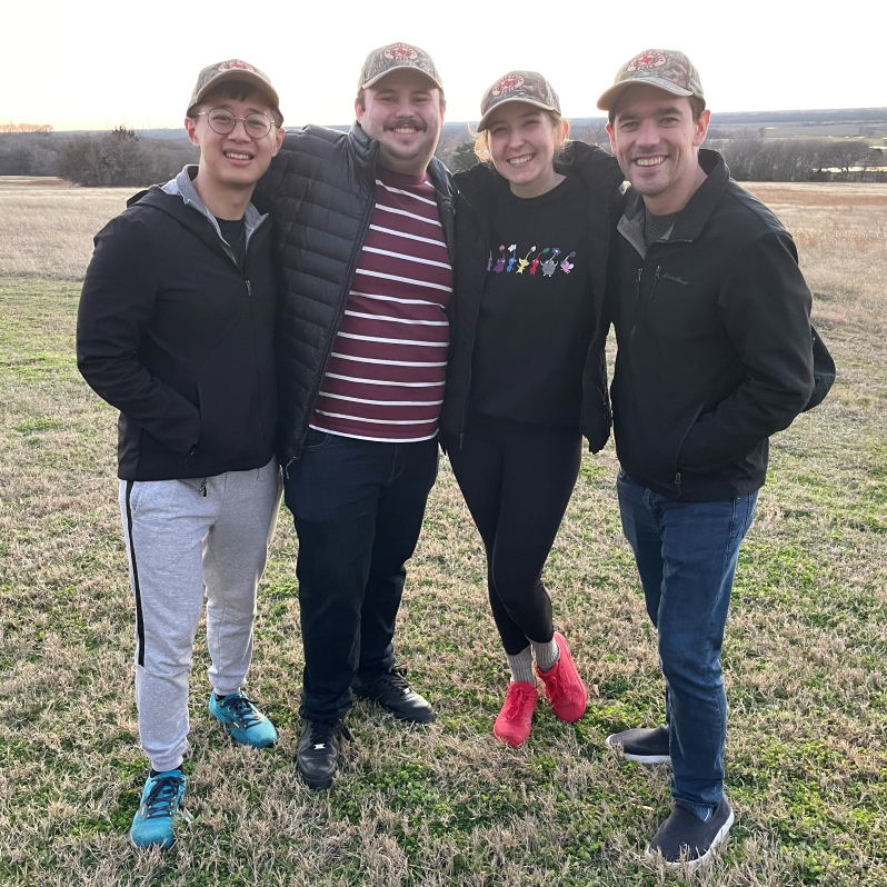 four people stand close together in grassy field, all wearing dark jackets and baseball caps