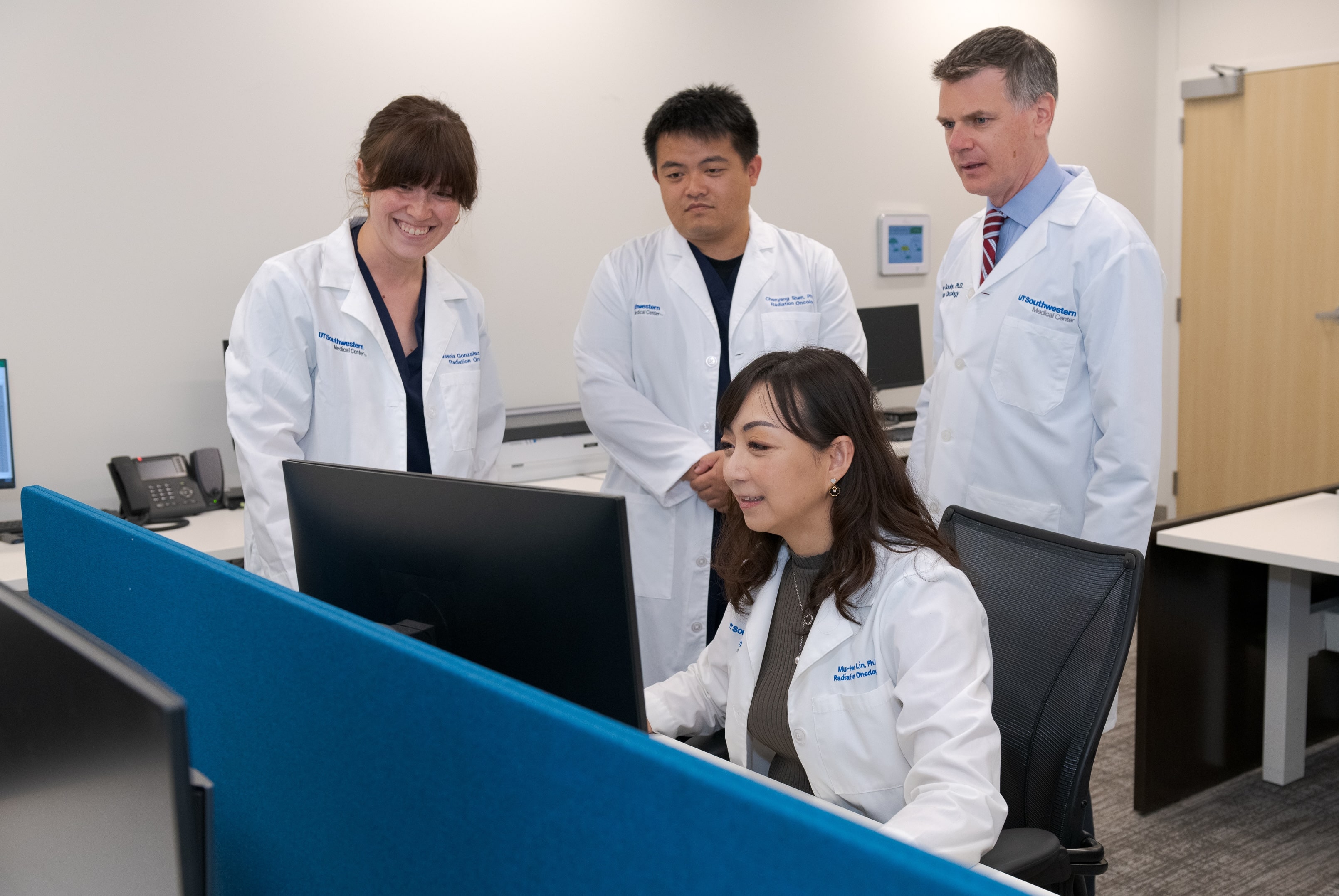 Four people in white lab coats look at a computer monitor.