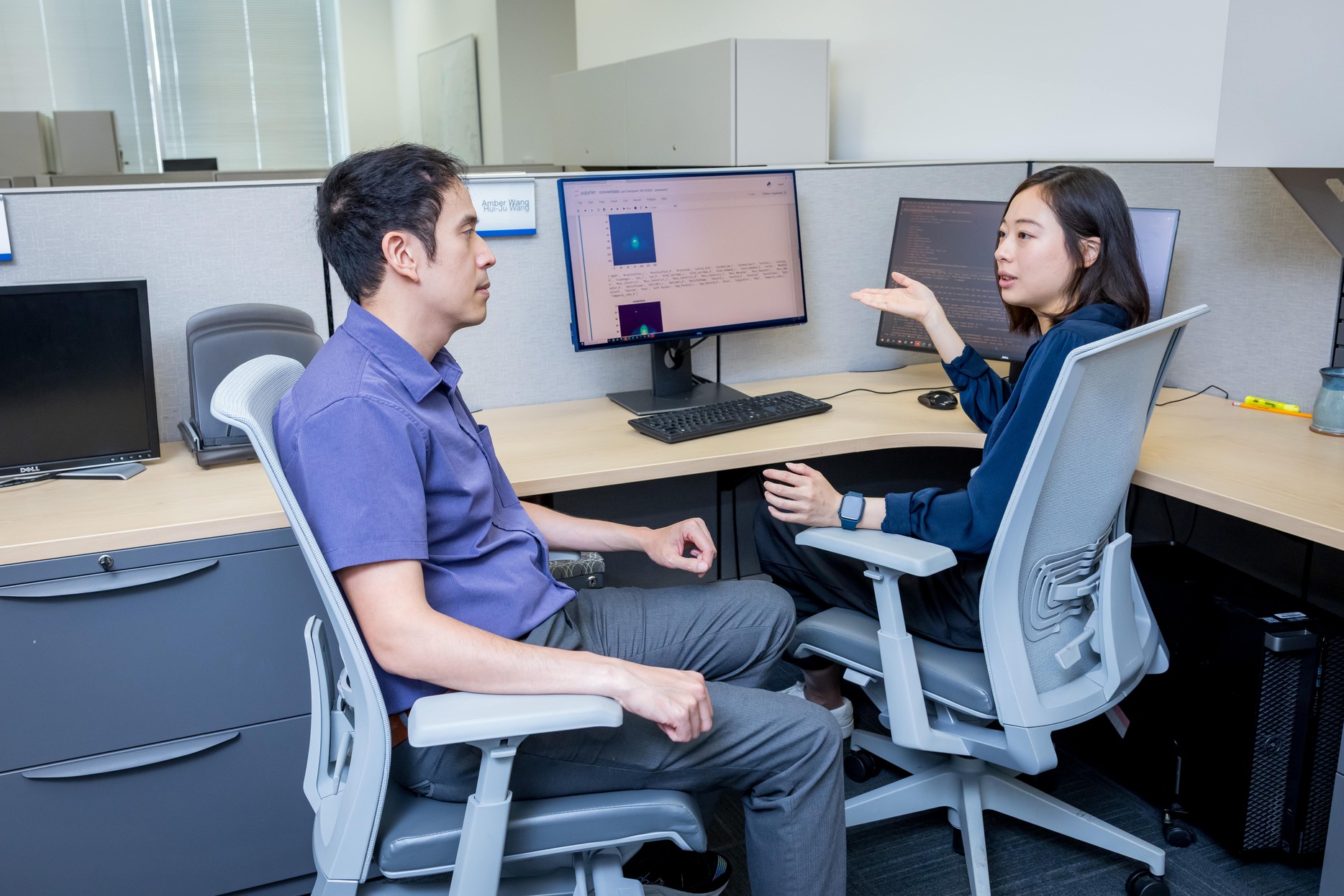 Two people sitting at a desk having a discussion with a monitor in the background.