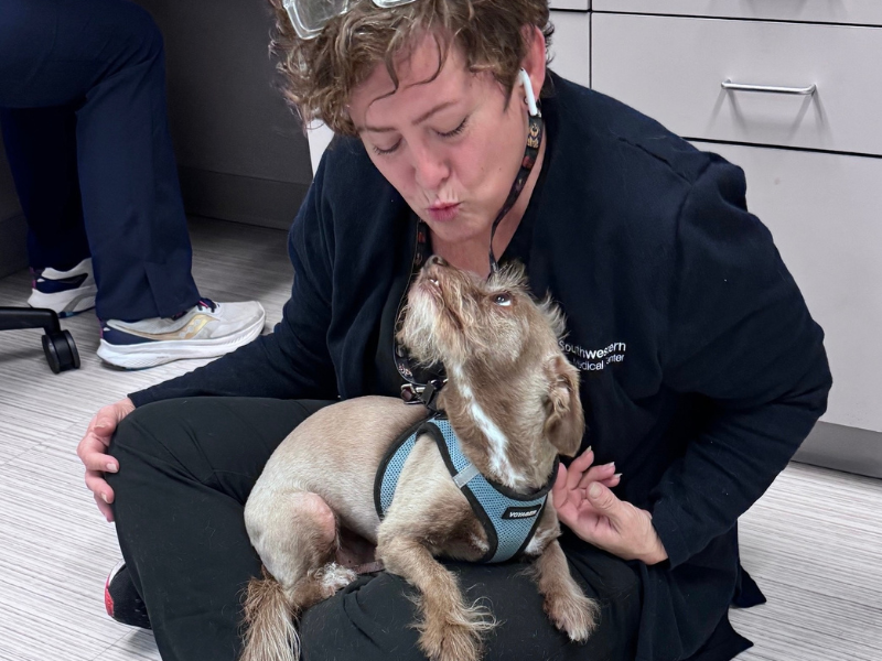 Woman in blue scrubs sitting down while holding a dog in her lap.