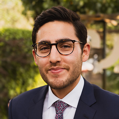 Smiling man with dark hair and dark facial hair, wearing a blue suit, white shirt, polk-dot tie, and dark rimmed glasses.