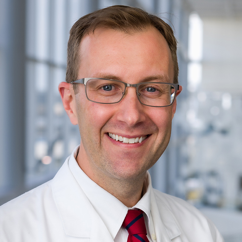 Smiling man with short brown hair, wearing a white lab coat, white shirt, red tie, and gray metal glasses.