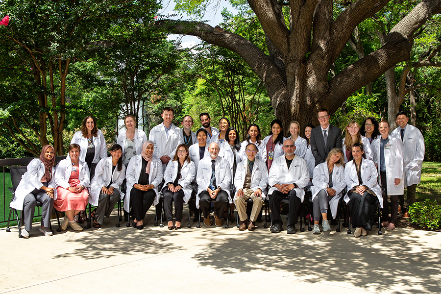 A group of smiling men and women wearing white lab coats, posed in front of a large tree.