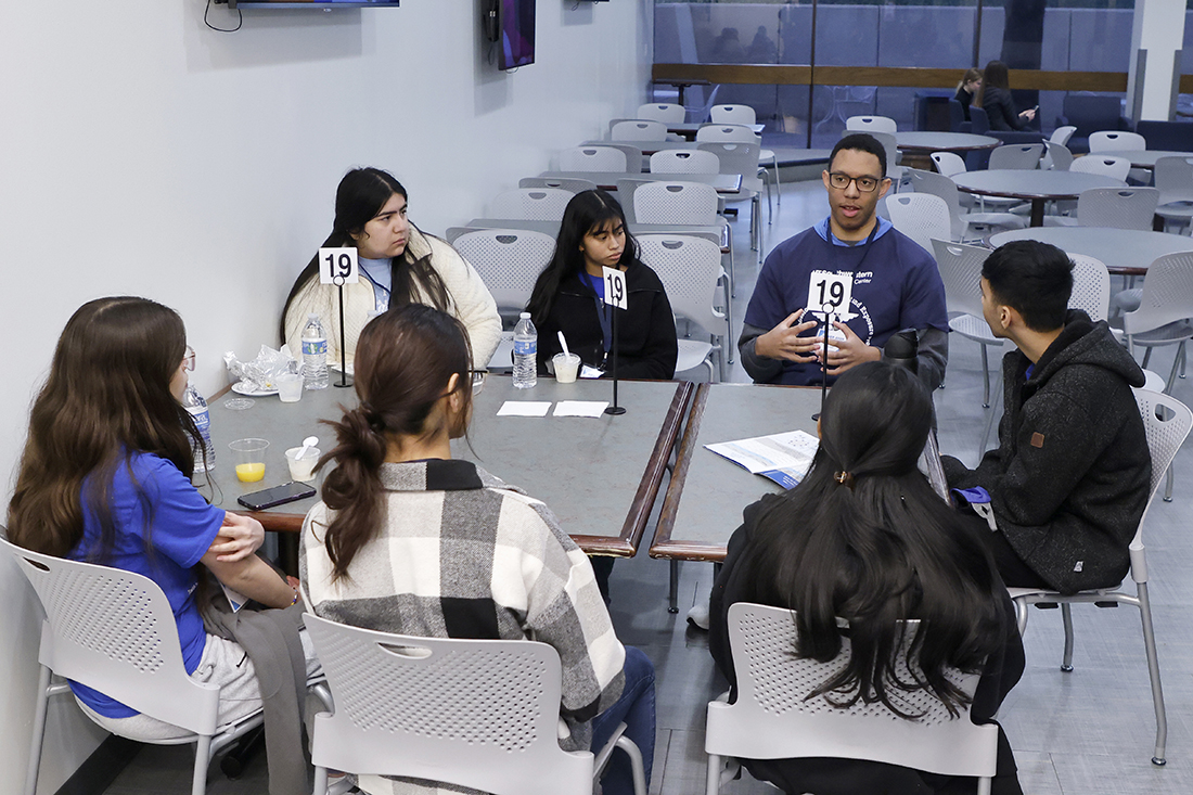 Six students sit at table with a male mentor who is speaking to them