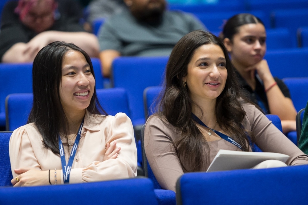 3 female undergraduate students sit in the audience (foreground) - from left, young Asian woman in pink shirt, woman with long dark hair in brown shirt, and in behind her woman with dark hair in a sleek bun wears a black shirt