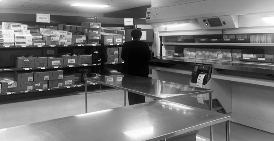 moody black and white image of person facing away in front of 2 walls of shelves full of medical drug boxes with empty metal exam table in the foreground