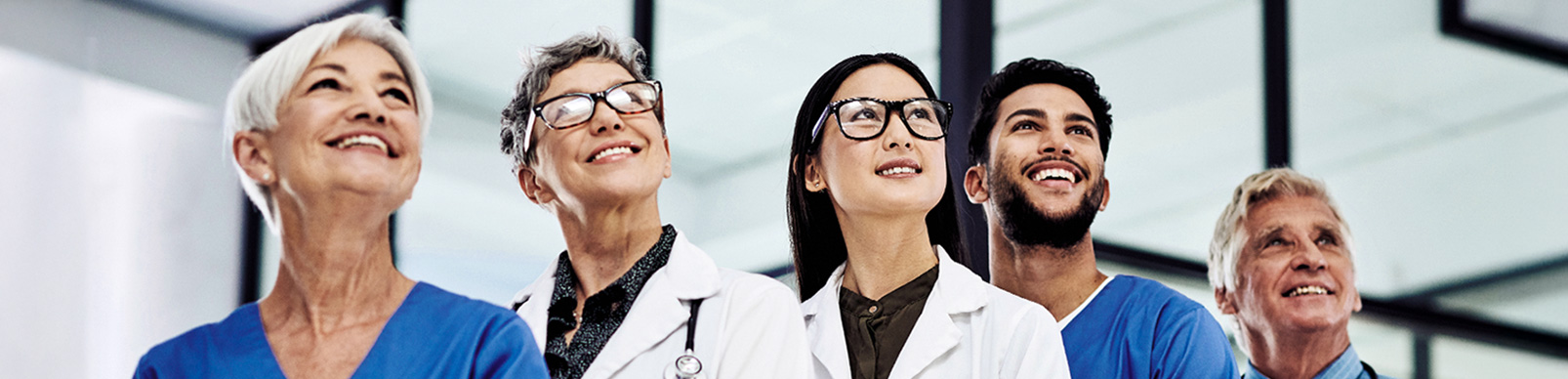 Three female and two male doctors in blue scrubs and white coats stand in a line looking up