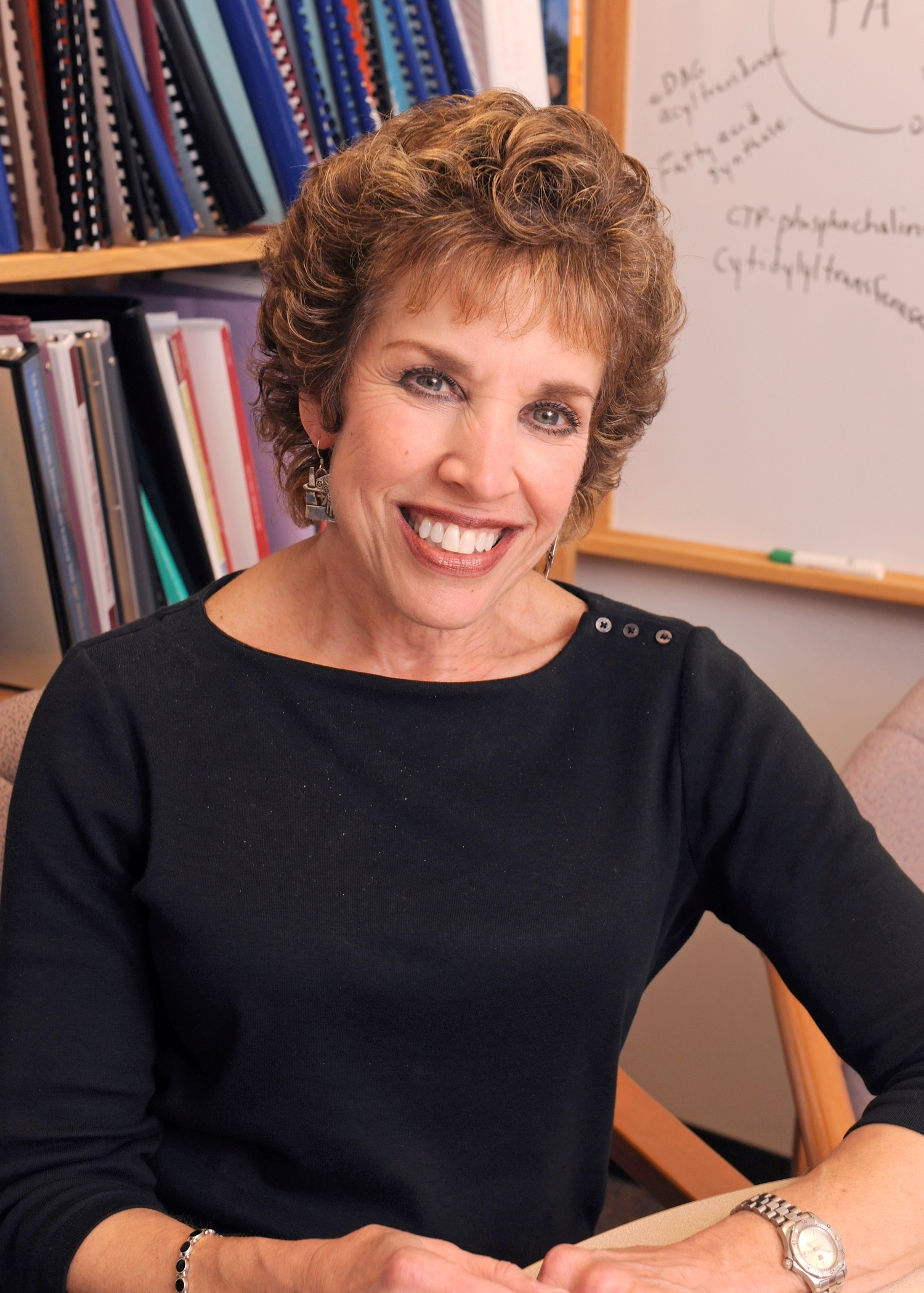 Smiling woman with short red curly hair, wearing a black sweater, seated at a desk.