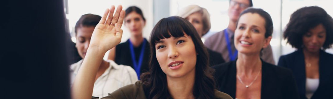 diverse group of professional adults seated in meeting, young woman with dark hair in center/front raises hand