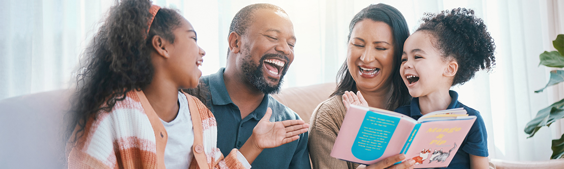 smiling family reading a children's book, black girl wearing orange striped sweater, black father, mother with long dark hair, younger girl with curly ponytail and blue shirt