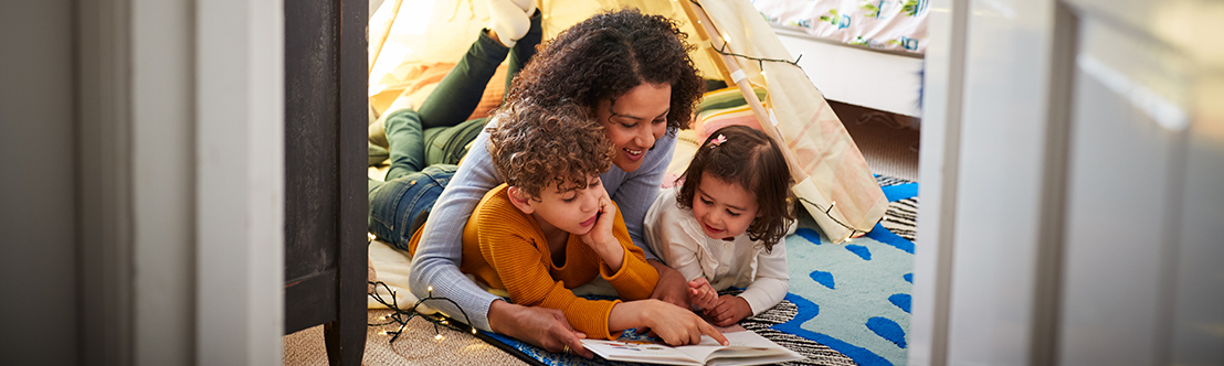 mother lies on floor under indoor tent reading with 2 children