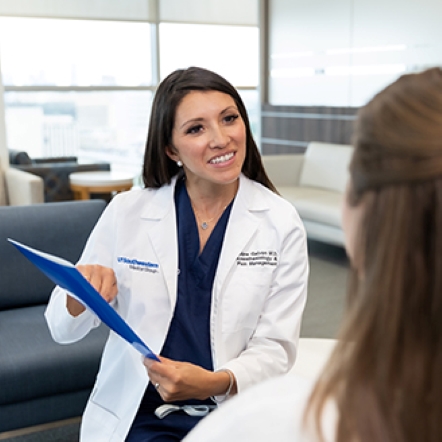 smiling woman with long dark hair wears lab coat and points to blue folder in her hand as she speaks to another woman in a hospital waiting area