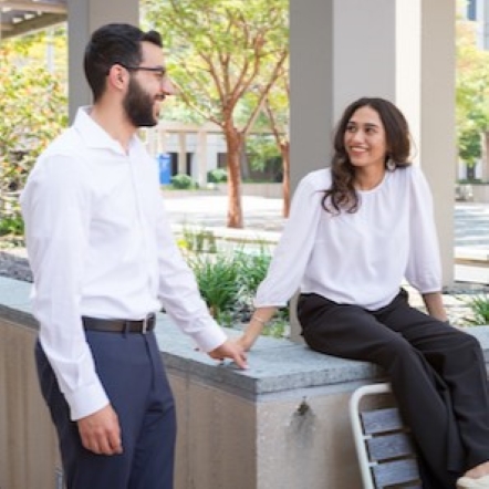 white male student with dark hair and beard stands outside on campus speaking to female student smiling and seated on a wall next to him