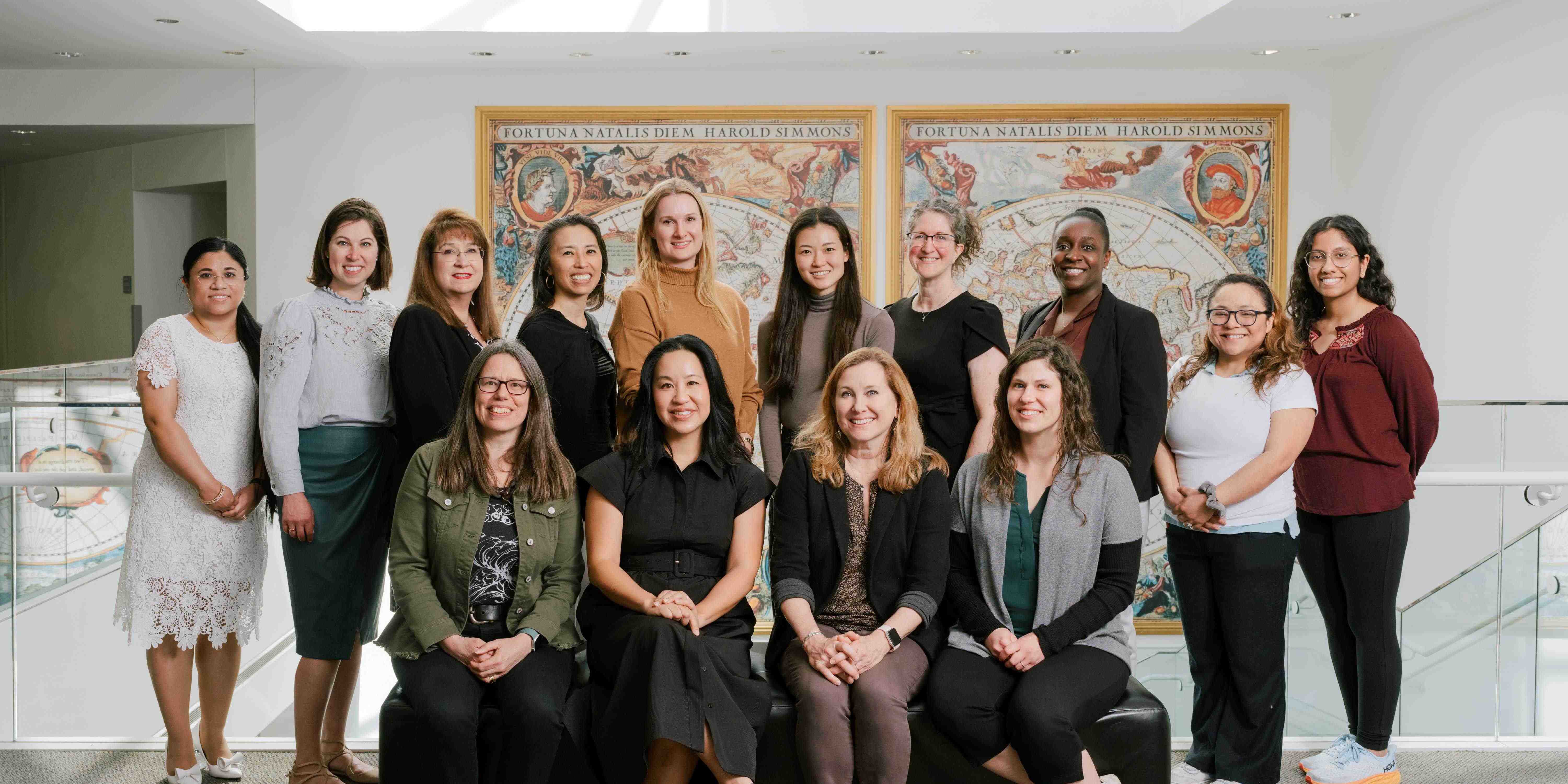 14 women, 4 seated and 10 standing, in front of a decorative painting of a world map.