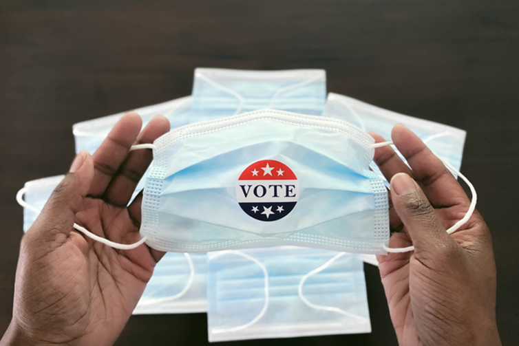 A Black physician holds a surgical mask with a Vote sticker on it