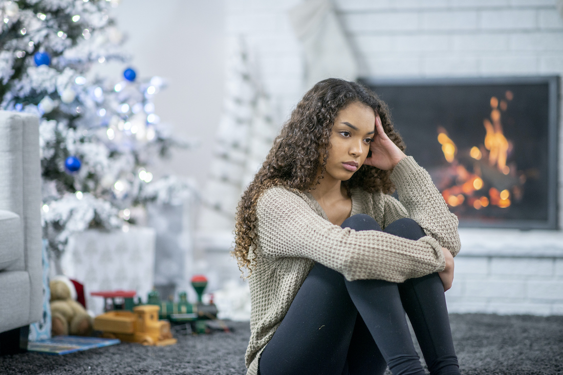 A girl sits on the carpet in her living room in front of the fireplace. She looks stressed and depressed as she sits alone on the holidays.