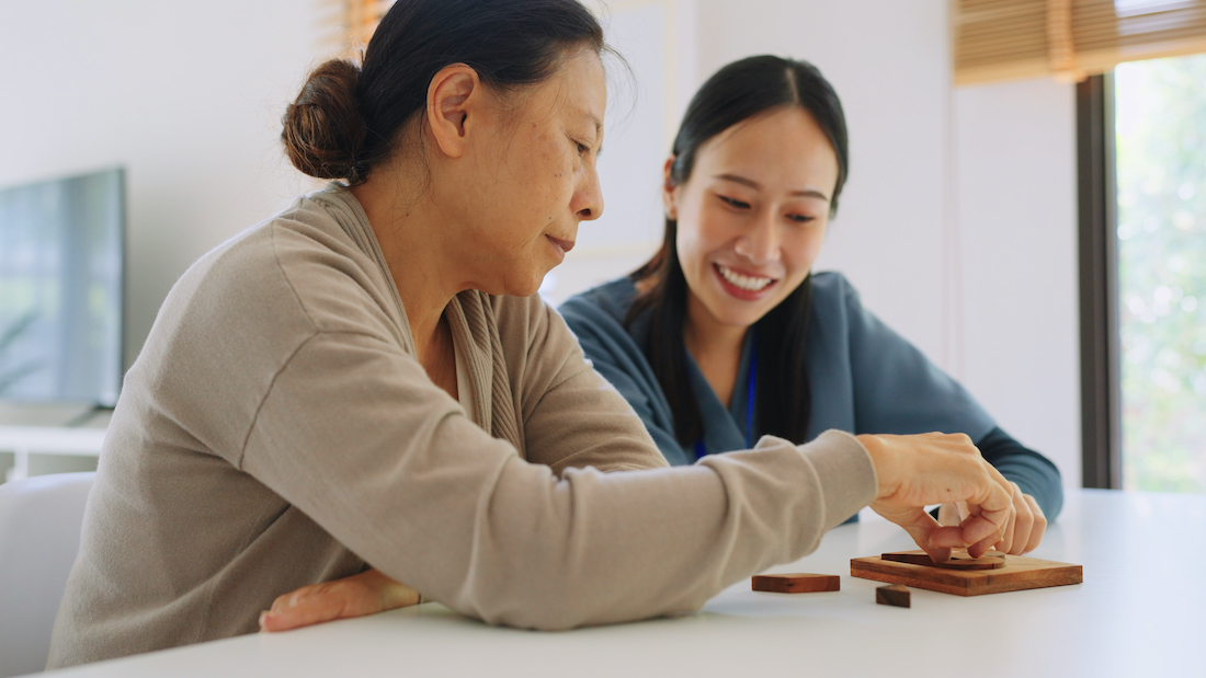 Caregiver nurse wearing blue scrubs and elderly woman retirement playing wooden shape puzzles game learning origami as for physiotherapy activity for dementia as a part of a physical therapy session medical health care plan