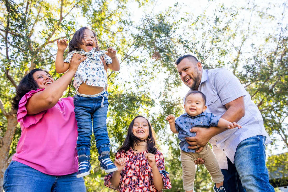 A family plays in the park, the parents lifting their smallest children up. The young boy has down's syndrome.