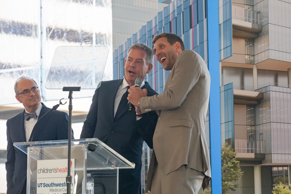 standing behind plexiglass podium, from left: balding man with glasses with suit and bow tie, holding microphone together - Troy Aikman man in gray suit and tie with Dirk Nowitzki taller man in tan suit